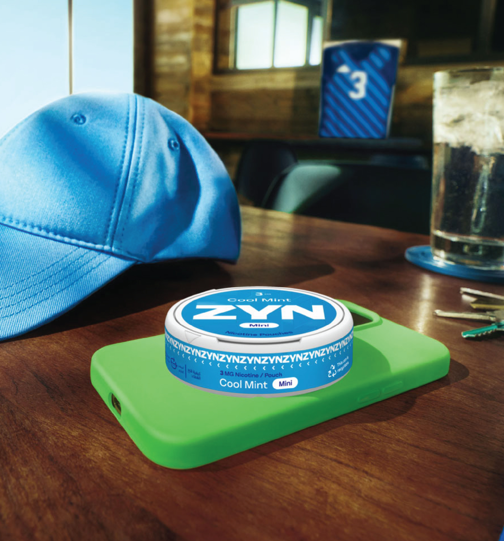 A ZYN container placed on top of a phone with a green case next to a blue cap, keys and a drink on a brown table